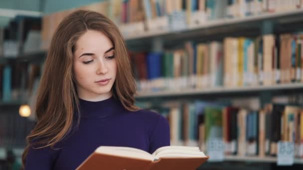 Задумчивая молодая студентка читает книгу, стоящую в библиотеке — стоковое видео