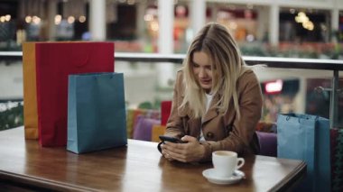 Mutlu sarışın kız alışveriş torbaları üstünde o ile kafede masada bir smartphone ile çalışır