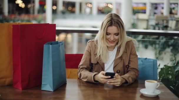 Chica rubia feliz trabaja con un teléfono inteligente en la mesa en el café con bolsas de compras en ella — Vídeos de Stock