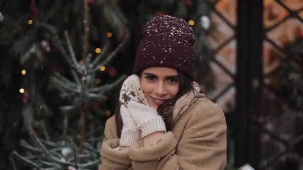 Close up di giovane carino felice bella ragazza in inverno vestiti in piedi in caduta fiocchi di neve, sorridente, tiene le mani vicino al viso a Natale decorato vetrina sfondo. Concetto vacanza invernale . — Video Stock