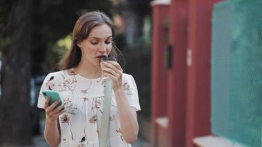 Beyaz çiçekli, çantasıyla çapraz giyinmiş, beyaz saçlı, beyaz saçlı, beyaz saçlı, güzel beyaz saçlı, akıllı telefonunu kullanan, Turkuaz Çitin yanında yürüyen kağıt bardaktan kahve içen bir kız..