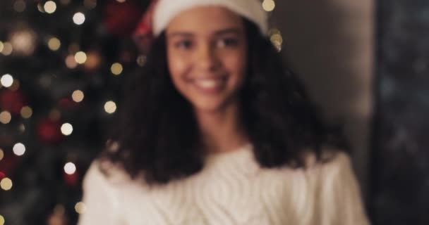 Young Smiling Mulatto Girl en Santas Hat le da la caja de regalo roja con cinta azul a la cámara, de pie en la iluminación del árbol de Navidad en el fondo. Feliz Concepto de Navidad. Primer plano . — Vídeo de stock