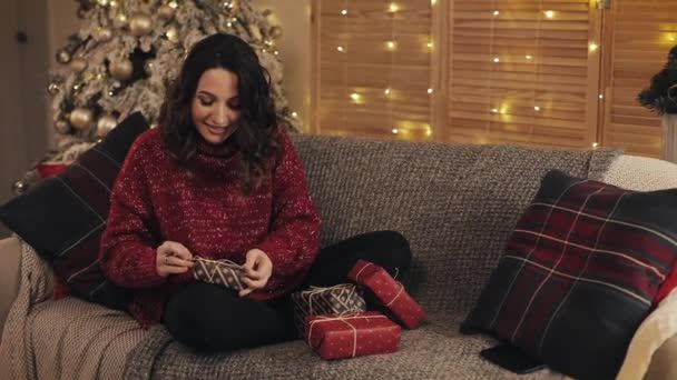 Piękne młode kobiety finalizuje prezenty świąteczne Taśma bandażowa i związany w łuk Siedząc na kanapie w pobliżu choinki w tle domu. Koncepcja wakacji i Nowego Roku. — Wideo stockowe