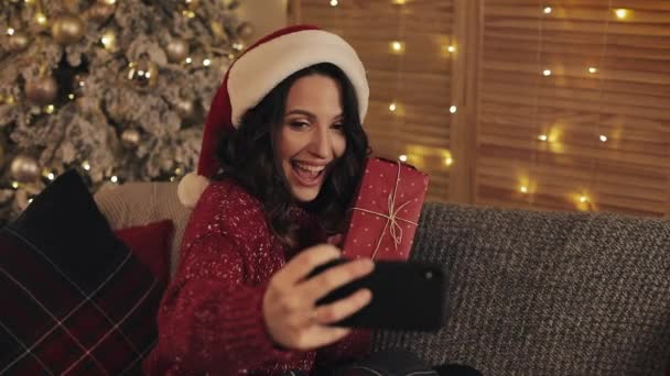 冬のセーターとサンタ帽子を身に着けている美しい若いブルネットの女性家の背景でクリスマスツリーの近くに座っている赤いプレゼント箱を持っているポーズセルフィーを作る。年末年始の概念. — ストック動画