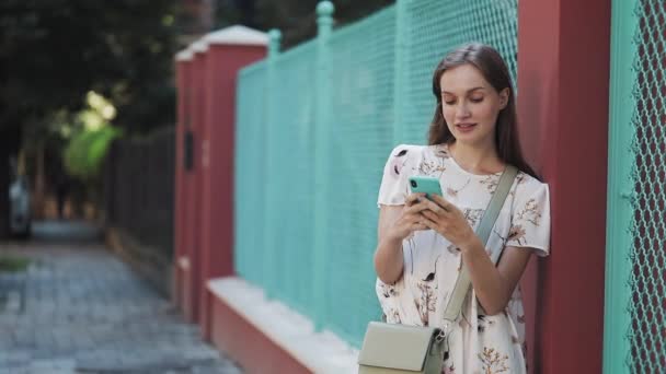 Joven Sonriente Encantadora Caucásica Chica Vestida de Flor Blanca y Bolsa Cruzó sus Hombros De Pie Apoyado en la Valla Usando su Teléfono Móvil Mirando a un lado Hablando Escribiendo . — Vídeos de Stock