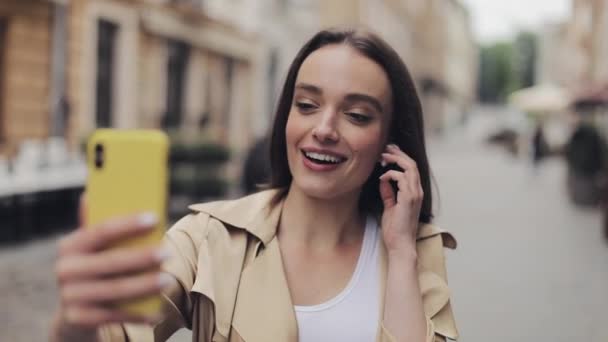 Pretty Usmívající se mladá dívka nosí sluchátka dotýkat se jich dělat videohovor držení telefonu vertikální mluvení a stojí na pozadí City Street Close Up. — Stock video