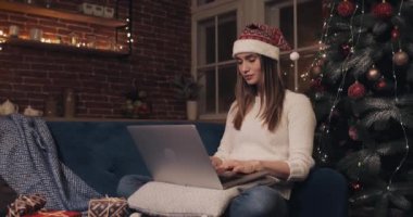 Noel Baba Şapkası Giyen Güzel Genç Kadın Ev Arkaplanı 'nda Noel ağacının yanında oturmuş Laptop Sohbeti Mesajlaşması Tatil ve Yeni Yıl Görünümü Konseptini kullanıyor..
