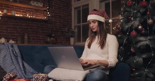 Красива молода жінка в капелюсі Сантас сидить біля Різдвяної ялинки на домашньому тлі Використовуючи її ноутбук балачки Концепція смс свят і новорічний вид збоку . — стокове відео
