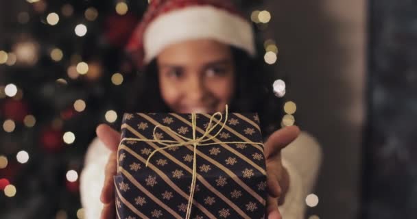 Pretty Smiling Mulatto Girl in Santas Hat Δίνει το χριστουγεννιάτικο δώρο κουτί στην κάμερα, στέκεται στο Διακοσμητικό Φωτισμό στο παρασκήνιο. Χειμερινές διακοπές Concept. Κοντινό πλάνο. — Αρχείο Βίντεο