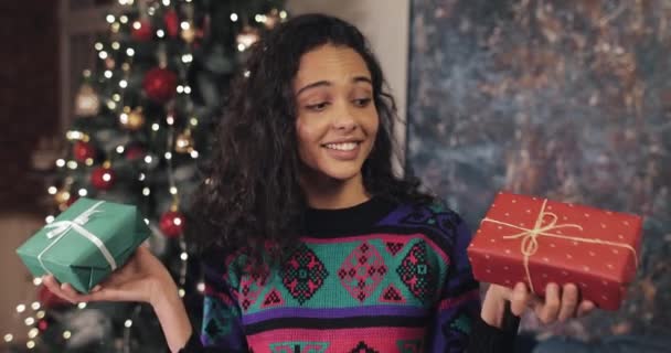 Mooi lachend meisje, met twee kerstcadeautjes in haar handen, schuddend, denkend welke te kiezen, Staande in de buurt van de kerstboom. Kerst en Nieuwjaar Feestconcept. — Stockvideo