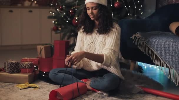 Attraente ragazza in Babbo Natale Cappello Preparazione regali di Natale, fasciatura nastro e legato in un arco, seduto sotto l'albero di Natale a casa Concetto di vacanze e Capodanno . — Video Stock