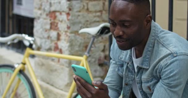 Fröhlicher afrikanisch-amerikanischer Typ, der mit seinem Smartphone ein Video anruft, winkt, lächelt, während er neben einem stylischen Fahrrad mit altem Metallzaun im Hintergrund sitzt. Seitenansicht. Nahaufnahme. — Stockvideo