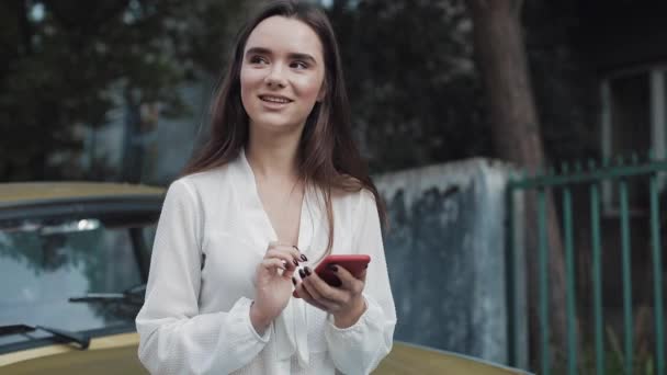 Jovem atraente sorridente morena caucasiano menina em pé inclinado para o velho elegante carro dourado ao usar seu telefone, conversando em redes sociais no Cosy Street Background . — Vídeo de Stock