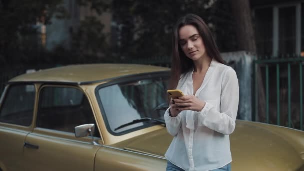 Joven atractiva morena caucásica chica de pie apoyado en el viejo coche de oro con estilo, mientras que el uso de su teléfono inteligente y pantalla de desplazamiento en acogedor fondo de la calle . — Vídeo de stock