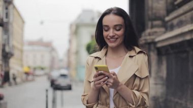 Young Preety Girl Holding Mobile Phone Texting and Laughing Standing at City Background .
