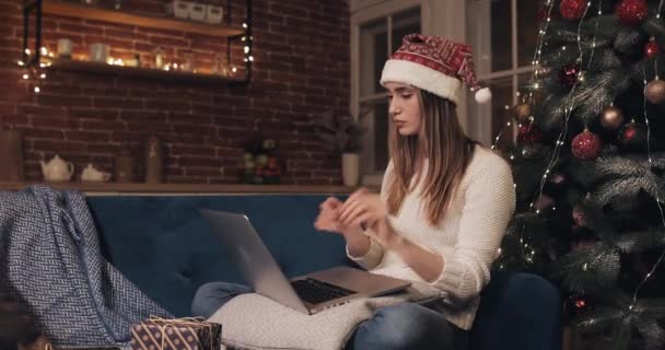 Attractive Caucasian Girl Sitting near Christmas Tree Using Her Laptop Thinking Looking at the Screen Ordering Present. Gift Box Falls into her Hands. Concept of Holidays and New Year Side View. — Stock Video