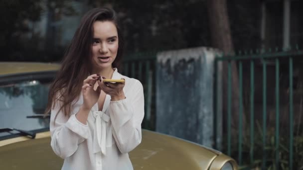 Menina branca bonita nova com cabelo marrom usando seu comandante da voz do smartphone ou fazendo a mensagem da voz ao estar inclinado ao carro dourado vintage na rua acolhedora . — Vídeo de Stock