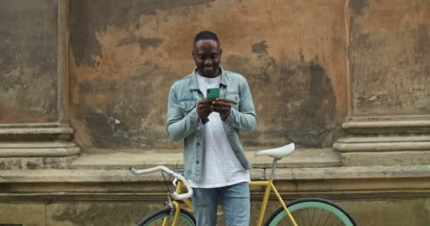 Jeune homme afro-américain portant des vêtements décontractés à l'aide d'un téléphone intelligent, regardant vers l'écran et souriant tout en se tenant penché sur le vieux mur du bâtiment en face du vélo moderne . — Video