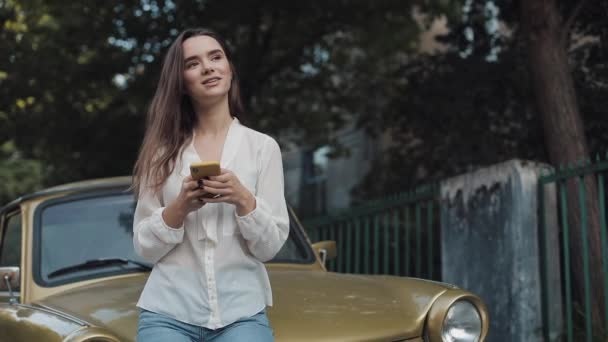 Jeune belle fille étudiante brune caucasienne utilisant l'application de téléphone portable et souriant tout en étant assis à Vintage Golden Car. Concept Technologie et Communication . — Video