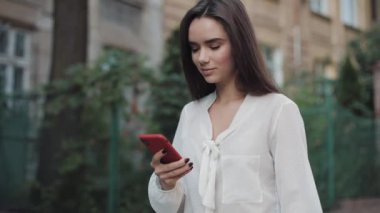 Güzel, gülümseyen esmer kız eski kasaba sokağında yürürken telefonunu kullanıyor ve etrafa bakıyor. İletişim ve Teknoloji Konsepti. Yan Görünüm