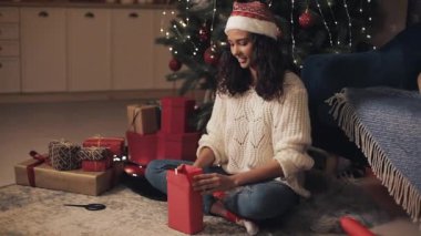 Noel Baba Şapkalı Çekici Mutlu Kadın Sunar, Şimdiyi Sarmak, Tatmin Olmuş ve Mutlu Görünmek, Konforlu Ev Arkaplanı 'nda Noel Ağacı Altında Oturuyor. Tatil Kutlama Konsepti.