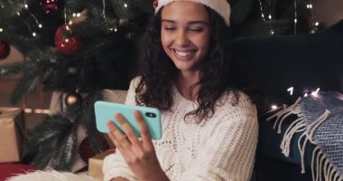 Noel Baba Şapkalı Gülümseyen Genç Mulatto Kız Yılbaşı Ağacı Altında Oturuyor ve Modern Cep telefonunu kullanarak bir video görüşmesi yapıyor, Gülüyor, Mutlu Görünüyor. İletişim ve Tatil Konsepti.