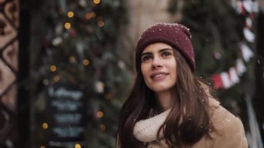 Kışlık Şapka Yürüyüşü Giyen Genç Beyaz Güzel Kız 'ın Kapanışı. Kar yağarken Yukarı Bakan Kadın Noel Dekorasyonlu Pencere Arkaplanda. Tatil Konsepti.