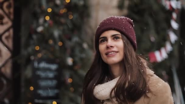 冬の帽子のウォーキングを身に着けている若い白人のプリティーガールの閉鎖。雪が降っている間の女性はクリスマスに見上げ窓の店の背景を飾る。. — ストック動画