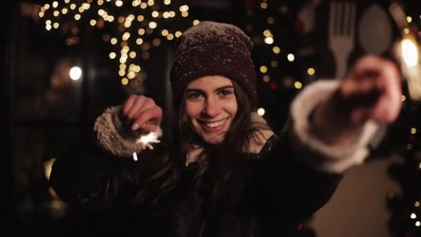 Pretty Smiling Girl en ropa de invierno, regocijo, mirando a la cámara. Mujer sosteniendo chispas y bailando, de pie en la caída de nieve al aire libre en Navidad decorado calle fondo . — Vídeos de Stock