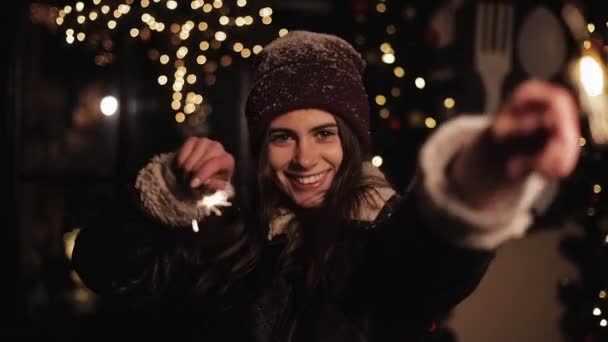 Bella ragazza sorridente in vestiti invernali, Rallegrandosi, Guardando alla macchina fotografica. Donna che tiene Sparklers e ballare, in piedi in caduta neve all'aperto in Natale decorato Street Background . — Video Stock