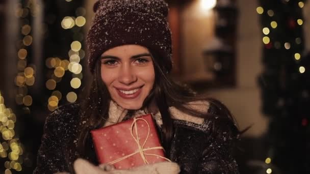 Nahaufnahme Porträt eines jungen lächelnden hübschen Mädchens, das glücklich aussieht, die Geschenkschachtel umarmt und lächelt. Frau steht im fallenden Schnee im Freien vor weihnachtlich geschmücktem Straßenhintergrund. Glückliches Urlaubskonzept. — Stockvideo