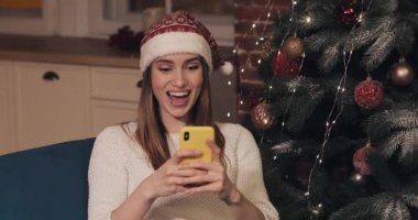 Noel ağacının yanında oturan, Noel Baba şapkası takan, akıllı telefonunu kullanarak 