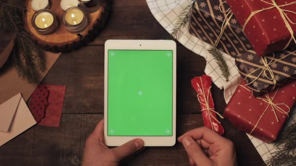 木製のテーブルの上のブランドの新しい白いタブレットの垂直緑の画面で手のスクロールとテーピングのトップビュークリスマスプレゼントとキャンドルがついています。モックアップ. — ストック動画