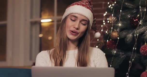 Усміхнена кавказька гарненька жінка у капелюсі Christamas сидить на дивані на затишному домашньому тлі біля різдвяної ялинки. Дівчина робить відео виклик за допомогою ноутбука Waving Привіт показуючи дерево — стокове відео