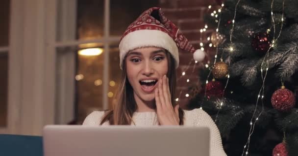 Mooi jong blond blank meisje dragen kerstmannen hoed zitten in de buurt van kerstboom met behulp van haar laptop chatten maken videocall zeggen Wow en glimlachen. — Stockvideo