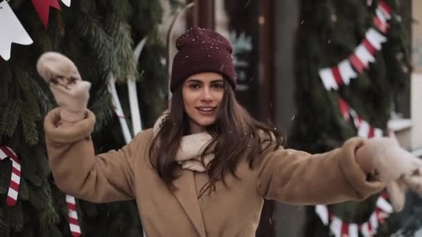 Молода щаслива чарівна дівчина в зимовому одязі стоїть в падаючих сніжинок зовні, посміхається, танцює, насолоджується погодою на Різдвяному прикрашеному тлі. Концепція зимового свята . — стокове відео