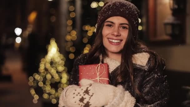 Primer plano Retrato de la joven y encantadora niña sonriente, luciendo feliz y satisfecha, sosteniendo la caja de regalo, de pie en la caída de nieve al aire libre en Navidad decorado fondo de la calle. Feliz concepto de vacaciones . — Vídeo de stock