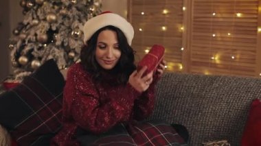 Noel Baba Şapkalı Çekici Esmer Kadın Portresi Grey Sofa Holding 'de Noel ağacının yanında oturuyor ve kameraya gülümseyen sallanan hediye kutusuna bakıyor..