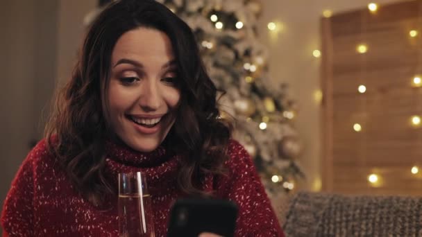 Close Up Ritratto di donna bruna attraente sorpresa Guardando il suo schermo dello smartphone e sorridendo. Ha in mano un bicchiere di champagne seduto a Accogliente sfondo casa vicino all'albero di Natale . — Video Stock