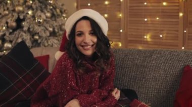 Noel Baba Şapkalı Güzel Esmer Kadın Noel Ağacı ve Parlak Işıklar Arkaplanı 'nın yanındaki koltukta oturuyor. Tatil ve yeni yıl kavramı. Kapat..