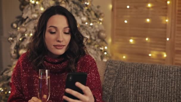 Hermosa mujer joven sosteniendo copa de champán sentada cerca del árbol de Navidad con luces brillantes en el fondo usando teléfono móvil y riéndose. Concepto de Vacaciones y Año Nuevo . — Vídeos de Stock