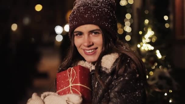 Закройте портрет молодой симпатичной девочки, выглядящей счастливой, держащей подарочную коробку и улыбающейся, стоящей в падающем снегу под открытым небом на фоне украшенной рождеством улицы. С праздником! . — стоковое видео