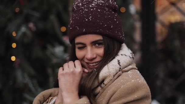 Primer plano Retrato de una joven con encanto usando sombrero de invierno, dando un guiño y sonriendo, mirando a la cámara, de pie en la ventana decorada de Navidad fondo de la tienda . — Vídeo de stock