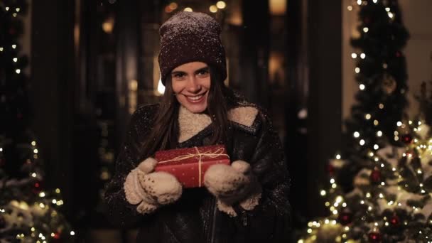 Happy Smiling Pretty Girl in Winter Clothes Holding Red Present Box, Looking Excited, Standing at Snowy Christmas Decorated City Street at the Background. Conceito de férias . — Vídeo de Stock