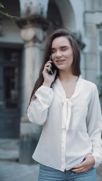 Hermosa joven sonriente morena chica hablando en su teléfono inteligente, de pie al aire libre en la vieja arquitectura en el fondo. Concepto de Comunicación y Tecnología. Vídeo vertical . — Vídeos de Stock