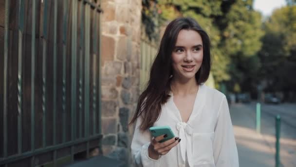 Mladá atraktivní usmívající se bruneta dívka kráčející po útulné ulici při použití svého telefonu a chatování v sociálních sítích s přáteli. Koncepce komunikace a techniky. — Stock video