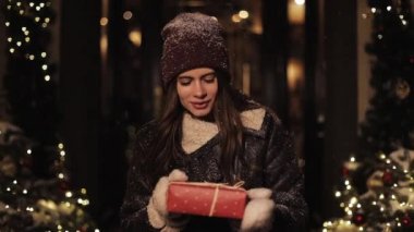 Mutlu Gülümseyen Güzel Kadın Kışlık Giysiler içinde Kırmızı Hediye Kutusu, tahmin et içinde ne var. Kız Kameraya Bakıyor Karlı Noel süslenmiş şehir sokağında duruyor