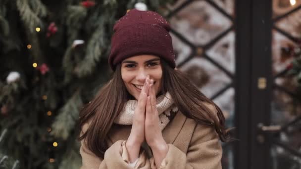 Retrato de una joven mujer sonriente con sombrero de invierno mirando a la cámara, calentando y frotando sus manos de pie afuera en el fondo de la tienda de ventanas decoradas. De cerca. Concepto de vacaciones . — Vídeos de Stock