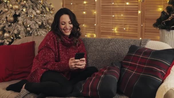 Hermosa joven morena sonriente en suéter rojo usando su teléfono inteligente escribiendo chat sentado cerca del árbol de Navidad en el sofá gris con cojines luces brillantes en el fondo interior — Vídeo de stock