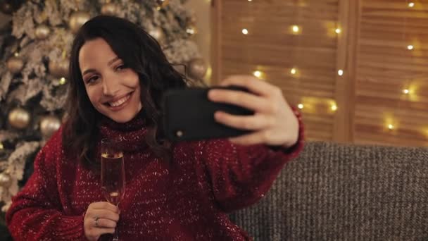 冬のセーターを着た魅力的な若いブルネットの女性がコスプレホームでクリスマスツリーの近くのソファに座っている背景シャンパングラスを保持するポーズセルフィーを作る — ストック動画